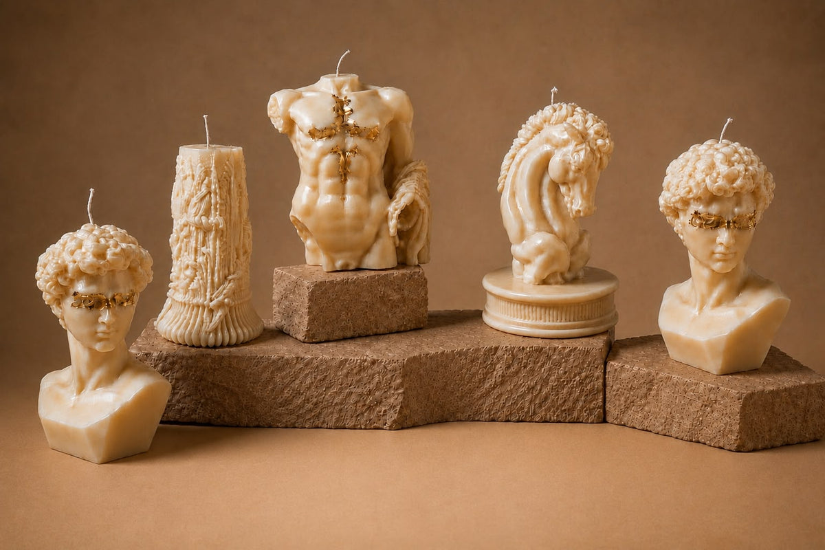 Five cream sculptural mould candles on textured stone plinths against a warm tan backdrop — a curly-haired classical bust, an ornate fluted column, a male torso with gold-leaf detail, a horse head and a second classical bust