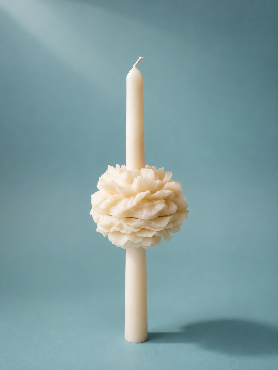 Slim cream taper candle with a sculpted peony bloom wrapped around its middle, standing upright against a teal backdrop